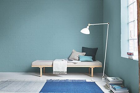 Gelassenes Mittelblau. Ein Raum mit einer in diesem harmonischen Farbton gestrichenen Backsteinwand und einer Liege davor.