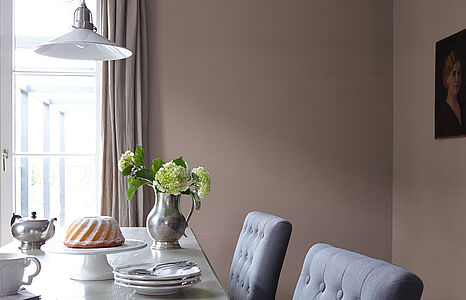 Nobles Erdbraun. Eine in diesem ruhigen und warmen Braun gestrichene Wand, davor ein Esstisch mit graublauen Polsterstühlen.