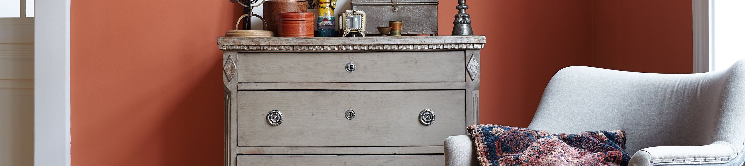 Eine Kommode in grauem Holz, ein grauer Sessel und Deko-Objekte vor einer Wand in gebranntem Rot.