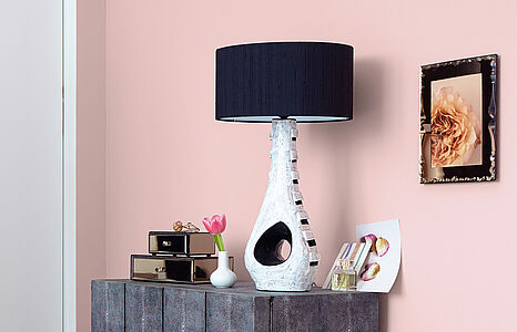 Verträumtes Graurosé. Ein Sideboard mit Lampe im Shabby Chic vor einer in diesem romantischen Farbton gestrichenen Wand.