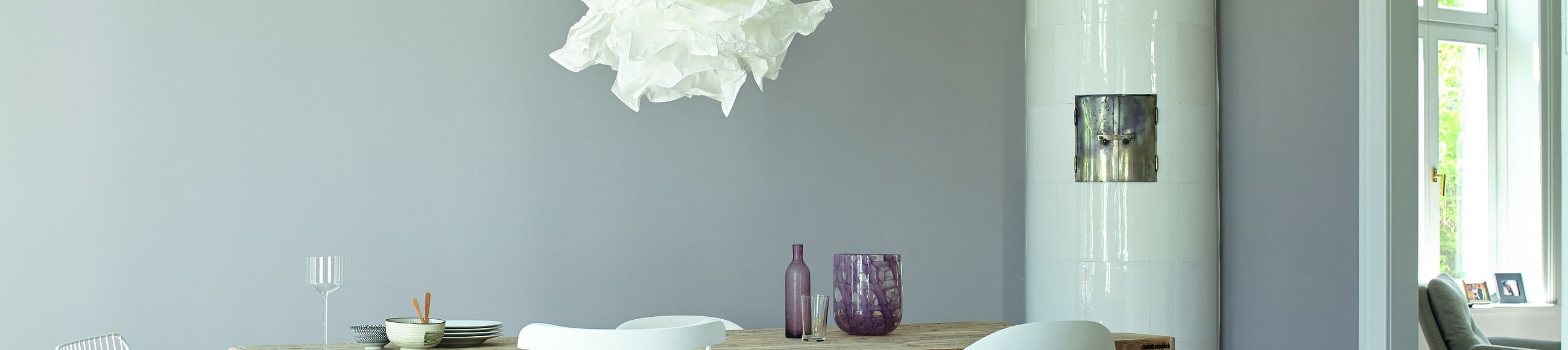 Großer Holztisch mit verschiedenen Stühlen und weißer Kamin vor hellgrauer Wand.