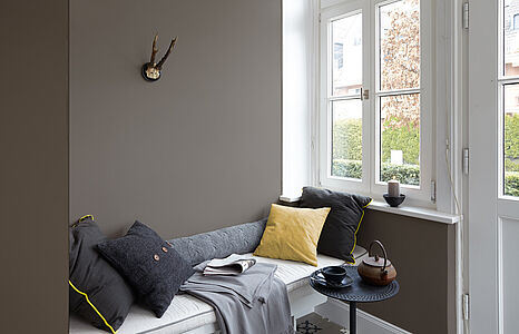 Zeitloses Grau. Ein in diesem behaglichen Farbton gestrichenes Wohnzimmer mit einer Liege mit Kissen unter einem Fenster.