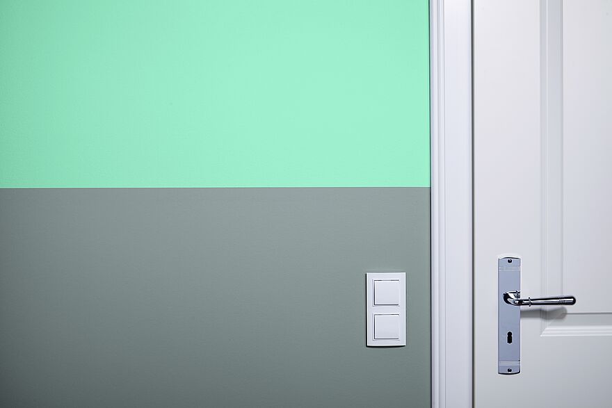 Eine zweifarbig gestrichene Wand mit einem grünen Farbstreifen oben und einem grauen Farbstreifen unten.
