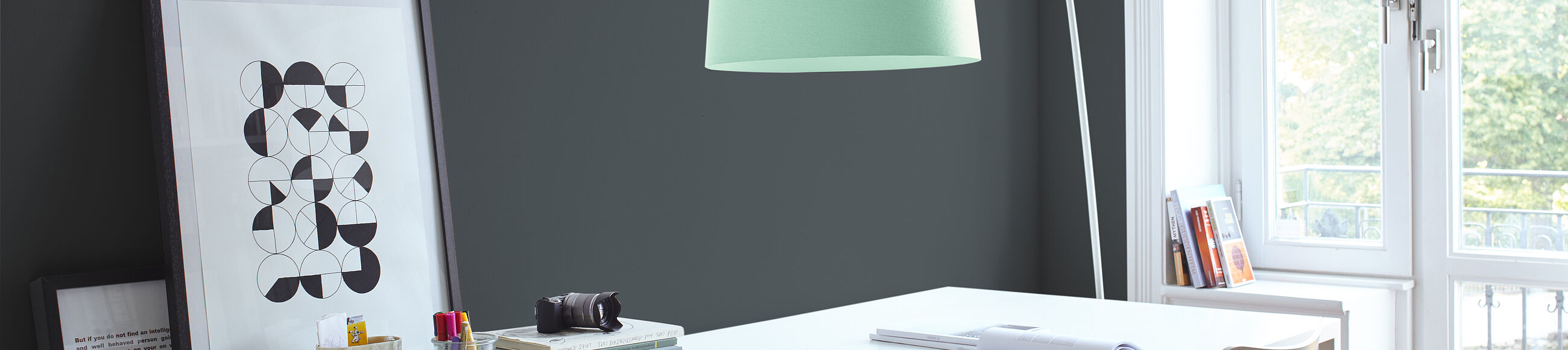 Ein weißer Schreibtisch vor einer dunkelgrauen Wand, daneben eine Stehlampe in Mint