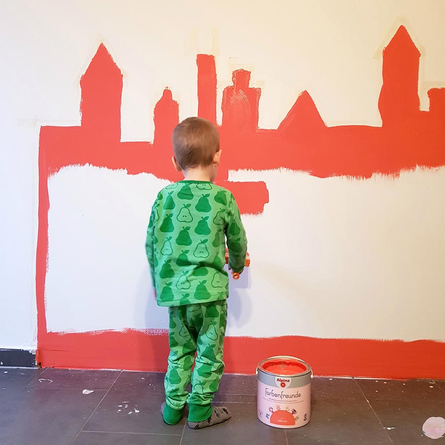 Eine Wand wird von einem kleinen Jungen mit Orange in Form einer Stadtsilouhette angemalt.