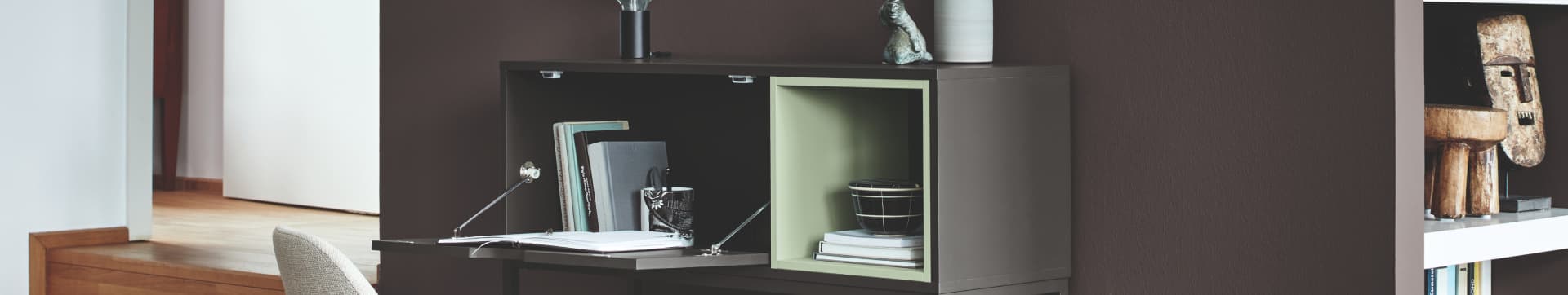 Ein dunkelgrauer, moderer, kastenförmiger Sekretär mit Schreibmaterialien, der vor einer in Dunkelgrau gestrichenen Wand steht.